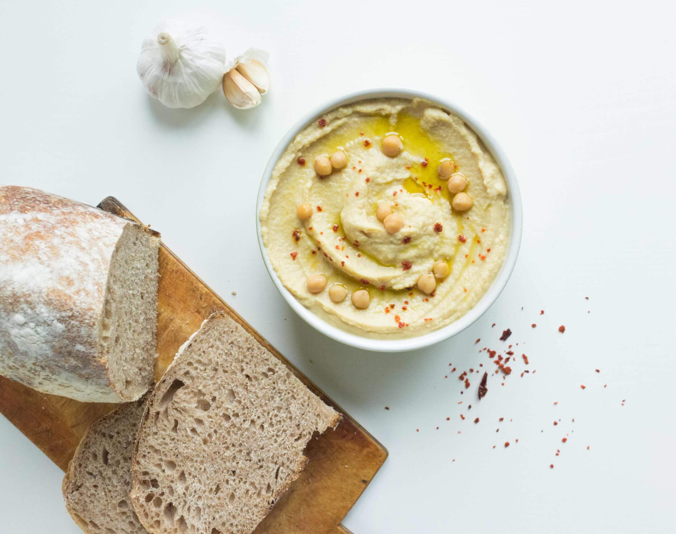 Eine weiße Schale mit Hummus daneben liegen ein Laib Brot, Chiliflocken und mehrere Knoblauchzehen.