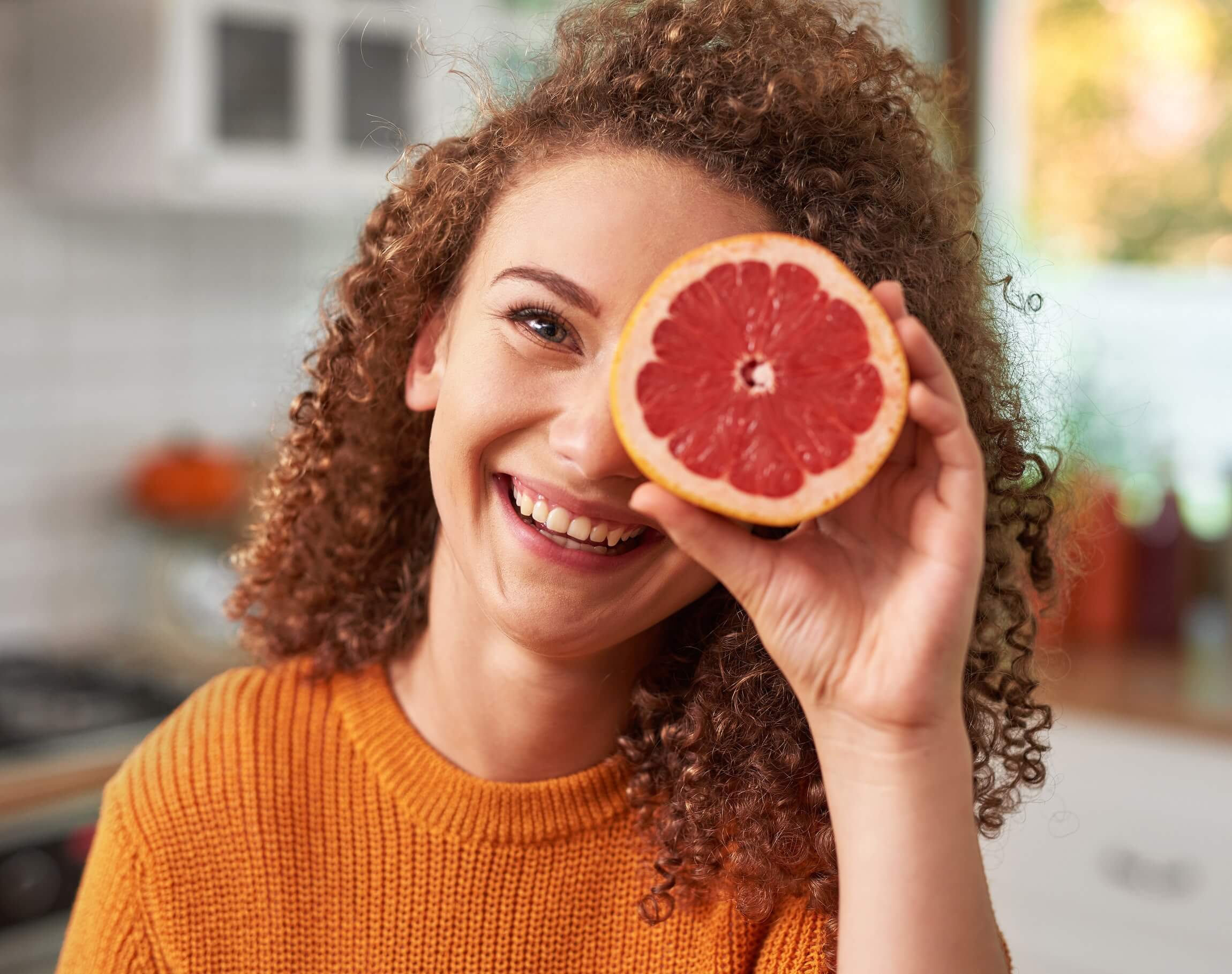 Junge Frau mit roten gelockten Haaren. Sie trägt einen orangen Pullover und hält sich eine halbe Grapefruite vor das linke Auge.