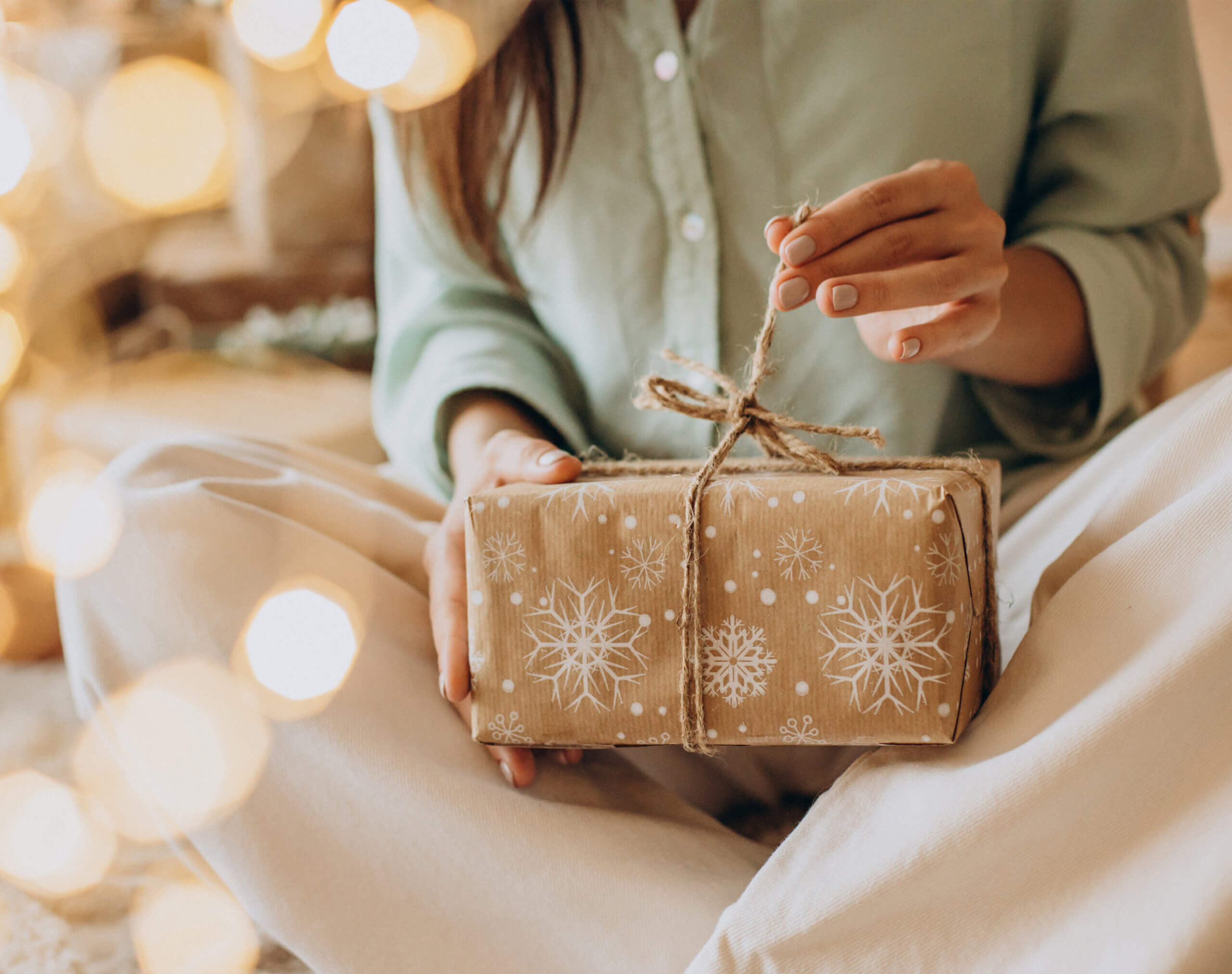 Eine Frau mit einer beigen Hose und einer hellblauen Bluse hält ein Weihnachtsgeschenk mit nachhaltiger Verpackung in der Hand.