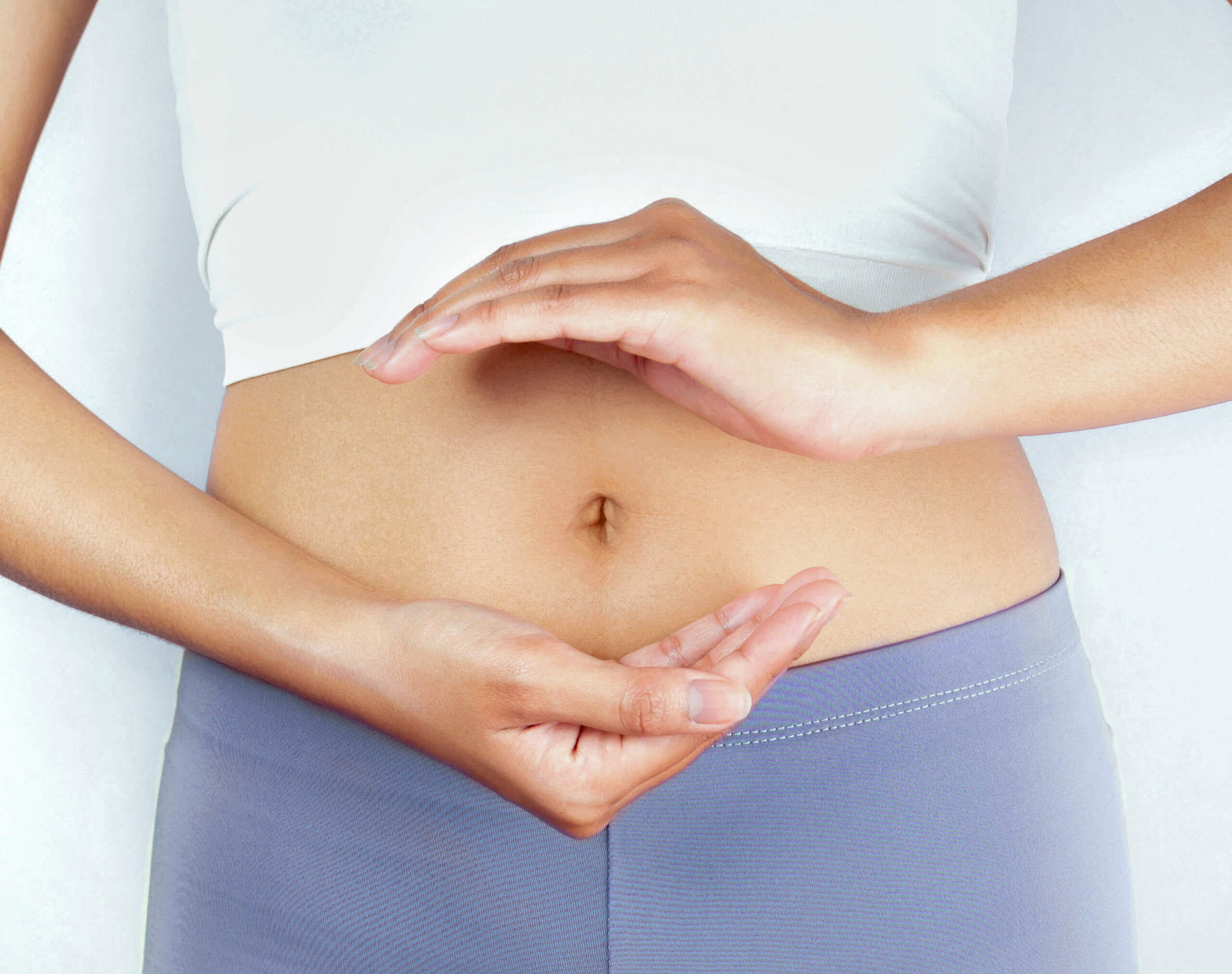 Frau die eine blaue Sporthose und ein weißes T-Shirt trägt und vor ihrem Bauch mit den Händen andeutet eine Kugel zu formen.